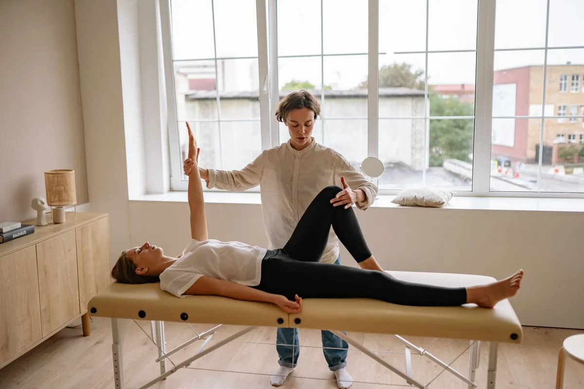 A physiotherapist guiding a patient's leg and arm stretch on a treatment table in a bright, window-lit room