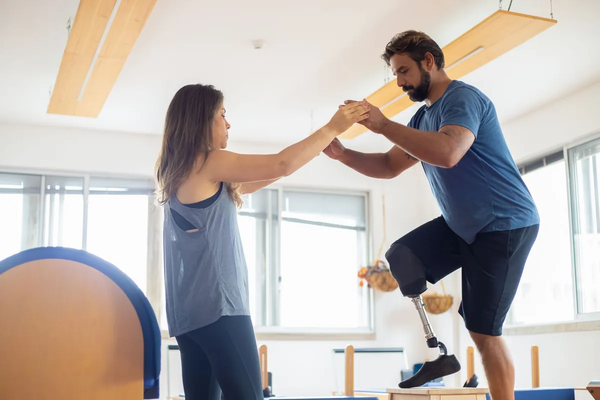 A physiotherapist steadying a patient with a prosthetic leg through a balance exercise in a bright rehab gym